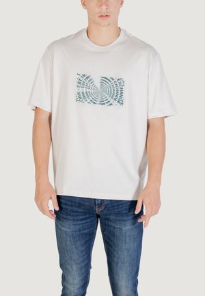 T-shirt bianco in cotone con un design grafico testuale circolare in teal. Maniche corte, vestibilità rilassata e collo a giro. Indossato con jeans blu scuro.