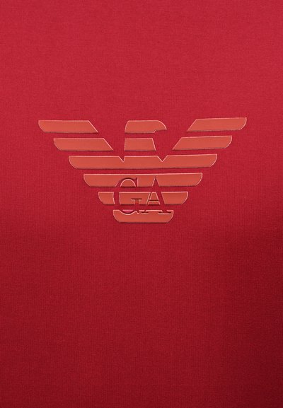 Κόκκινο ύφασμα με ένα γυαλιστερό, στυλιζαρισμένο λογότυπο αετού που διαθέτει οριζόντιες ρίγες και τα γράμματα "GA" στο κέντρο.