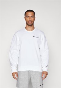 Sweatshirt blanc à col rond en coton avec des manches longues, arborant un petit logo noir et rouge sur la poitrine gauche. Texture douce, coupe décontractée.