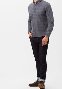 Szara koszula w kratę z długim rękawem i guzikami, noszona z ciemnymi dżinsami i brązowymi zamszowymi butami. Koszula ma klasyczny kołnierz i dopasowany krój.