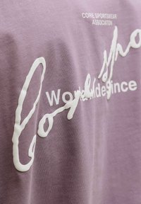 Levandų spalvos audinys su iškilusiais baltais užrašais „Core Sportswear Association“ ir „World wide since longshore“ mišriomis šrifto stilistikomis.