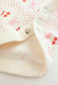 Cream-colored baby onesie with pink and red cherry and bow patterns, featuring metal snap buttons on a ribbed trim.