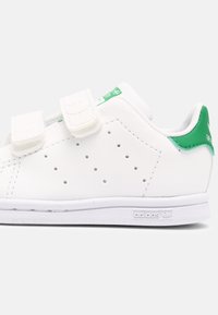 White leather sneaker with green accents, twin Velcro straps, perforations on the side, and textured white rubber sole. Adidas logo on heel.