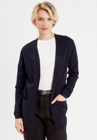 Cárdigan azul marino de abertura frontal, con dos bolsillos delanteros y una tela texturada. Se combina con una camiseta blanca y jeans oscuros.