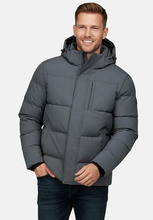 Man glimlachend met een grijze gewatteerde winterjas met kap, handen in de zakken, staand tegen een effen witte achtergrond.