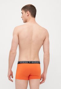 Shorts de boxe orange avec une ceinture noire portant le logo "GUESS". Le tissu semble lisse et ajusté. Le modèle est montré de dos.