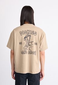 Trumpas smėlio spalvos marškinėliai trumpomis rankovėmis, ant nugaros esantis žaismingas grafikas su tekstu „DICKIES“ ir „LAND AHOY!“ paryškintu juodu šriftu.