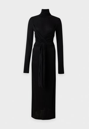 Femme aux longs cheveux foncés portant une robe noire à manches longues avec col roulé et un détail portefeuille, ainsi que des talons ouverts noirs, debout devant un fond uni.
