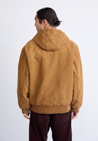 Homme avec de longs cheveux en chignon portant une veste à capuche marron et un pantalon marron foncé, tourné vers un fond clair uni.