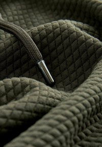 Mørkegrøn stof med en tekstureret, diamantmønstret overflade. Har et flettet brunt snoretræk og en metallisk aglet.
