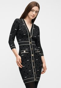 Γυναίκα με ίσια καστανά μαλλιά, ντυμένη με στενή μαύρη φόρεμα με λευκά γεωμετρικά σχέδια και χρυσά κουμπιά, ποζάρει μπροστά σε ένα απλό φόντο.