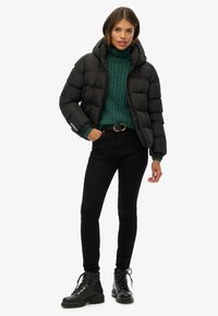 Svart puffjacka med huva, bärs över en grön kabelstickad tröja, ihop med svarta skinny jeans och svarta snörkängor.