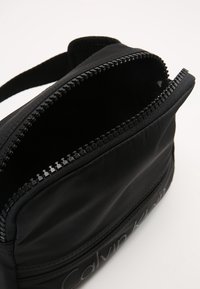 Schwarze Nylon-Tasche mit strukturierter Oberfläche, ausgestattet mit einem Reißverschluss oben und einem eingeprägten Logo, das ein kompaktes und funktionales Design bietet.