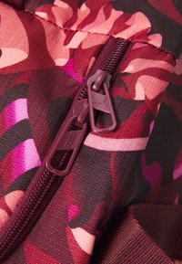 Close-up van een maroon, roze en zwart gemusterd stof met een dubbele rits en een klein Puma-logo op een van de ritszweepjes.