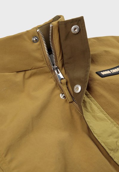 Khakifarbener Jackenkragen mit einem metallischen Reißverschluss, Druckknöpfen und einem kontrastierenden hellgrünen Innenfutter. Mit einem Logopatch auf der Vorderseite.