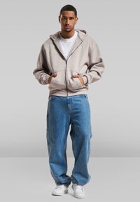 Beige zip-upsjakke med frontlommer og snorhette, kombinert med løse blå denimjeans og hvite joggesko. Teksturert stoff, avslappet passform.
