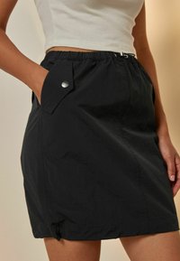 Jupe noire, légère, avec une taille élastique, une poche latérale avec fermeture à pression et un ourlet légèrement volanté. Texture lisse.