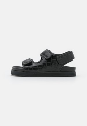 Sandalias negras con textura grabada tipo cocodrilo, correas ajustables de velcro, parte trasera abierta y suela gruesa de goma negra.