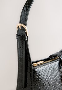 Sac à main noir à motif crocodile avec une finition brillante, doté d'une boucle dorée sur la sangle réglable et d'un compartiment zippé.