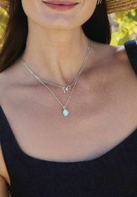 Silverfärgade lagerhalsband har en liten rosettcharm och ett hänge med en blå sten. Bäraren har en naturlig hudton och mörka kläder.