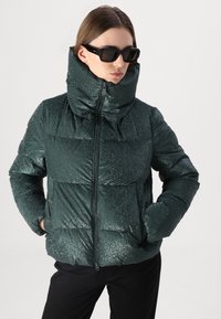 Groene pufferjack met een glanzende, getextureerde afwerking, hoge kraag, ritsluiting en zijzakken. Zwarte zonnebril maakt de look af.