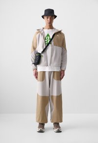 Light grey and tan fleece zip-up jacket with oversized pockets, paired with wide tan pants. Black bucket hat, white T-shirt with green print, and sneakers.