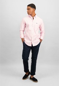 Camicia button-up rosa chiaro con un piccolo logo nero, abbinata a pantaloni chinos blu navy e espadrillas nere, in piedi contro uno sfondo neutro.