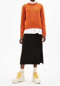 Personne portant un pull en maille orange par-dessus une chemise blanche, une jupe midi noire avec une fente sur le côté, des baskets montantes crème avec des lacets jaunes.