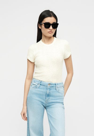 Γυναίκα με μακριά σκούρα μαλλιά που φοράει μαύρα γυαλιά ηλίου, λευκό κοντομάνικο πουκάμισο και ανοιχτό μπλε τζιν ψηλόμεσο, όρθια με το ένα χέρι στην τσέπη.