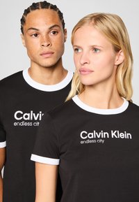 Juodos marškinėliai su baltais rausvais apykaklėmis, ant priekinės dalies atspausdinta "Calvin Klein endless city" kontrastingomis spalvomis. Abu drabužiai yra prigludę.