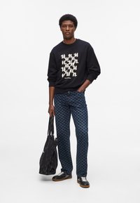 Čierna mikina s krémovými grafickými vzormi, modré džínsy s houndstooth vzorom, čierna taška a čierne tenisky s bielymi šnúrkami a gumovými podrážkami.