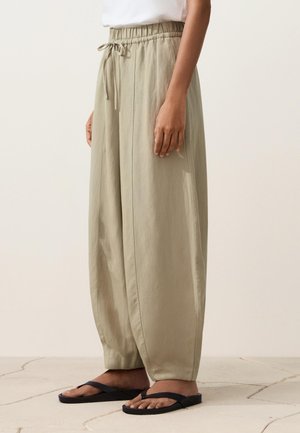 Személy laza, bézs színű, megkötős nadrágot és fekete papucsot visel, sima háttér előtt csempézett padlón áll.