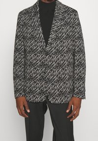 Blazer a motivi bianco e nero con un design a onde testurizzato, colletto con revers e chiusura a due bottoni, indossato sopra un dolcevita nero.