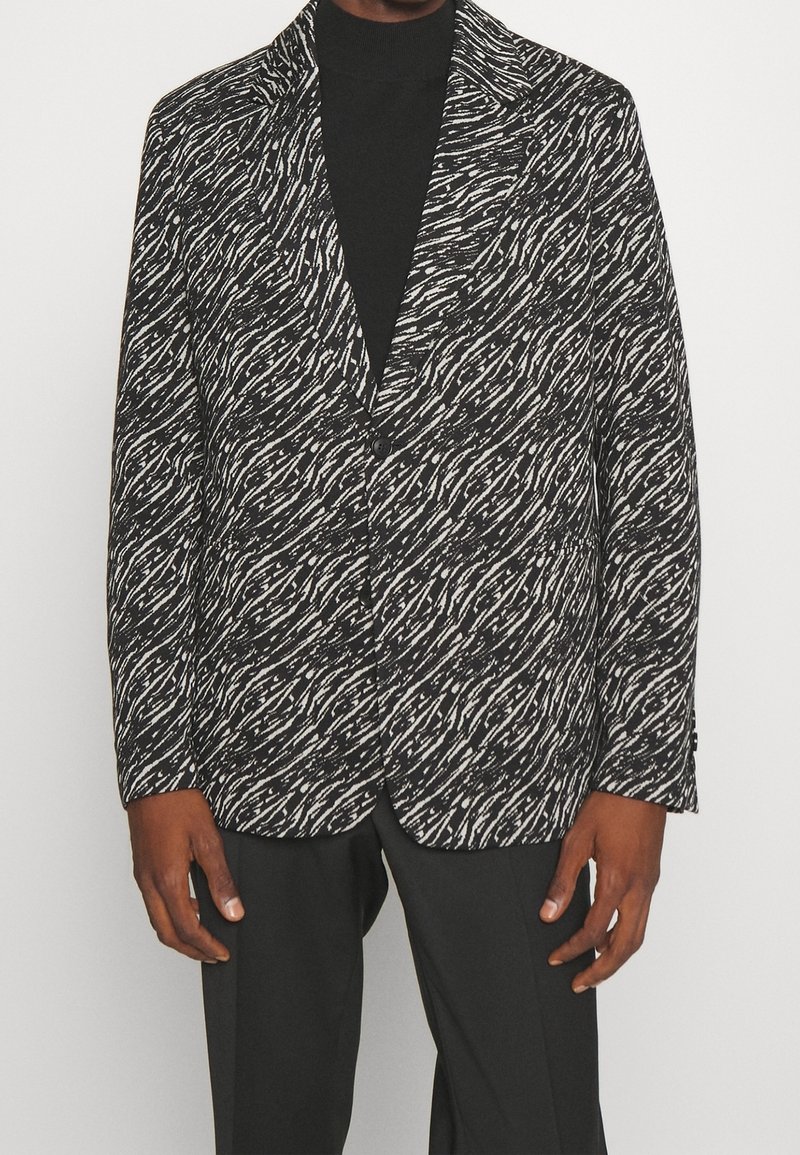 Blazer a motivi bianco e nero con un design a onde testurizzato, colletto con revers e chiusura a due bottoni, indossato sopra un dolcevita nero.