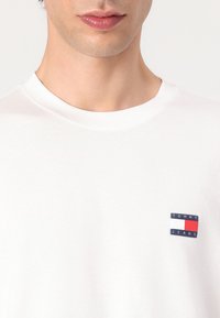 Witte katoenen t-shirt met een klein logo-opdruk in marineblauw, rood en wit op de linkerborst, met een ronde hals en korte mouwen.