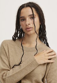 Pull en maille beige avec un col rond, des poignets côtelés et une texture douce. Des mèches de cheveux longs tressés entourent le visage.