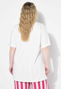Bílý oversized tričko s ohrnutými rukávy, s hladkou texturou. Model má na sobě pruhované růžové kalhoty. Pohled zezadu zdůrazňuje délku.