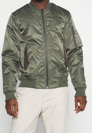 Uomo che indossa una giacca bomber verde con cerniera frontale, polsini e orlo a coste, tasca con cerniera sulla manica e pantaloni beige.