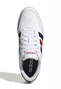 Bijela atletska cipela s kožnom gornjom dijelu, vezanjem na vezice, crvenim i tamnoplavim prugama, te logotipom na peti. Jastučić na jeziku i mrežasti podstavljeni materijal za udobnost.