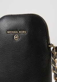 Musta muotopahvista valmistettu käsilaukku, jossa on kultaiset yksityiskohdat, "Michael Kors" -logo ja ketjureunus, sekä vetoketjukiinnitys.