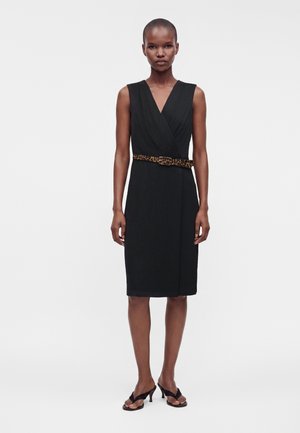 Femme portant une robe noire sans manches longueur genoux avec un col en V et une ceinture imprimé léopard, associée à des sandales noires à talons.
