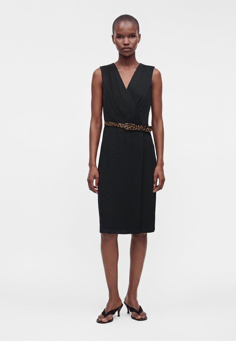 Femme portant une robe noire sans manches longueur genoux avec un col en V et une ceinture imprimé léopard, associée à des sandales noires à talons.