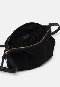 Borsa a tracolla nera in suede con cerniera dorata parzialmente aperta, che mostra l'interno vuoto e la tracolla in pelle regolabile su sfondo bianco.