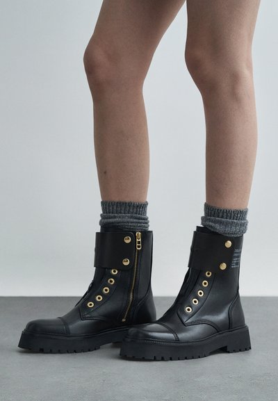 Black leather combat boots with gold hardware, side zipper, lacing detail, and a thick, textured sole. Features fold-over cuff design.