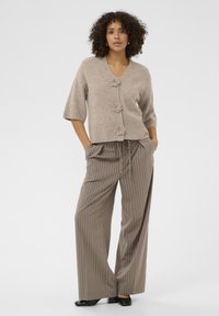 Beige cardigan with three floral buttons, short sleeves, and ribbed hem. Paired with loose, brown pinstriped pants and black shoes.