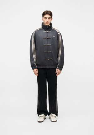 Jeune homme portant une veste Adidas foncée avec fermetures à cordon, un pantalon noir assorti avec des bandes, et des baskets Adidas blanches, debout devant un fond blanc.