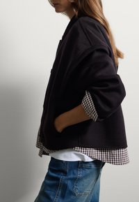 Veste en mélange de laine noire au look décontracté, dotée d'un col haut et de fermetures à boutons, superposée à une chemise à carreaux marron et blanc.