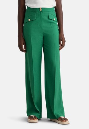 Femme grande portant un pantalon vert taille haute à jambes larges avec des boutons dorés et des poches à rabats sur le devant, associé à des sandales beiges.