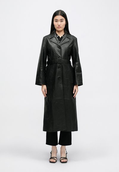 Cappotto trench in pelle nera con vita a cintura, maniche lunghe e chiusura a bottoni. La texture è liscia con cuciture visibili e tasche frontali.