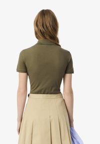 Polo shirt verde oliva con colletto, maniche corte e finitura testurizzata, abbinato a una gonna beige plissettata. Vista posteriore mostrata.